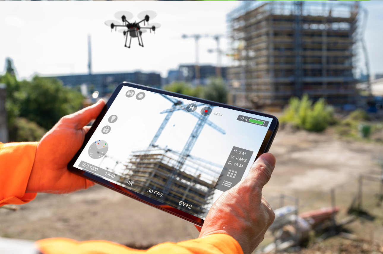 Two hands holding an electronic tablet in the foreground while a drone inspects a large industrial construction project in the background.